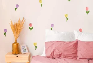 Koselig barnerom med veggdekor av blomster i pastellfarger. Sengen har rosa sengetøy og puter, mens en vase med tørkede blomster og ramme med bilder står på nattbordet.