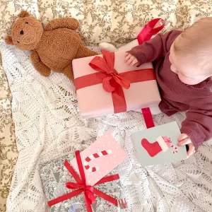 Baby, der sidder og kigger på julegaver med røde sløjfer og julekort på.