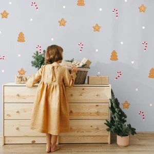 Vægsdekoration med julemotiv af peberkager formet som juletræer og stjerner samt candy canes, med en lille pige i kjole som står foran en kommode.