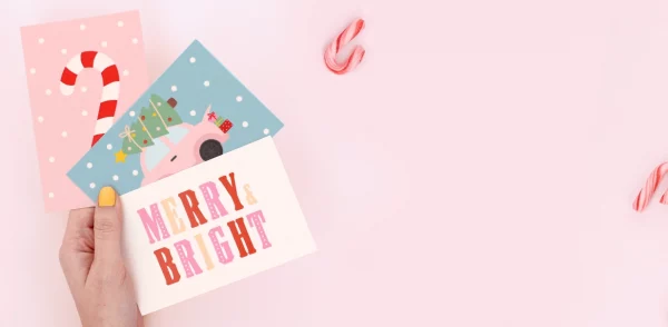 Hånd der holder farverige fødselsdagskort med julemotiver og teksterne “Merry Bright” og “Ho Ho Ho” på en julegave.