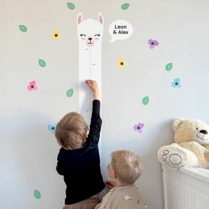 Børn måler sig på en vægdekoration med lama-motiv, omgivet af farverige blomsterklistermærker. Perfekt til børneværelset.