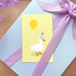 Børnefødselsdagskort med en illustreret and med gul ballon og festhat, placeret på gaveæske med lilla bånd, ideel til børnefødselsdage.