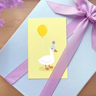 Billede af en farverig fødselsdagskort med en tegning af en and, der bærer en gul ballon. Kortet ligger ovenpå en gave indpakket med lilla bånd, perfekt til børnefødselsdage. Køb unikke kort til barnerummet og festlige lejligheder hos os