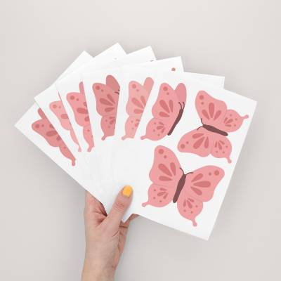 Hånd, der holder ark med vægklistermærker med rosa sommerfugledesign, perfekt til børneværelser.