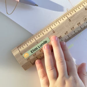 Billede af en hånd, der holder en trælineal med en navnelap, der viser navnet Elias Lunde. Lappen har et sødt design med en løve. Baggrunden indeholder tegnematerialer og papir, hvilket indikerer kreativ leg. Dette billede illustrerer brugen af navnelapper, der er perfekte til at personalisere børns ejendele.