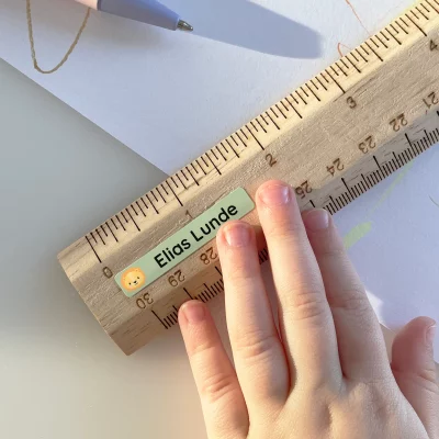 Billede af en hånd, der holder en trælineal med en navnelap, der viser navnet Elias Lunde. Lappen har et sødt design med en løve. Baggrunden indeholder tegnematerialer og papir, hvilket indikerer kreativ leg. Dette billede illustrerer brugen af navnelapper, der er perfekte til at personalisere børns ejendele.