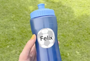 En håndholdt vandflaske med navnet Felix påklæbet en fodboldmotiveret navnelap. Flasken er blå og står på en grønn græsplæne, hvilket giver et aktivt og udendørs tema. Perfekt til sport og leg for børn.