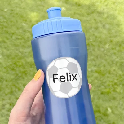 En håndholdt vandflaske med navnet Felix påklæbet en fodboldmotiveret navnelap. Flasken er blå og står på en grønn græsplæne, hvilket giver et aktivt og udendørs tema. Perfekt til sport og leg for børn.