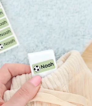 Navnelap med navnet Noah synlig i en hånd, der holder et stykke tøj med en grøn sportstema fotball. Ved siden af ligger et par grønne børnesko og et blødt tæppe. Perfekt til at personliggøre børns ejendele.