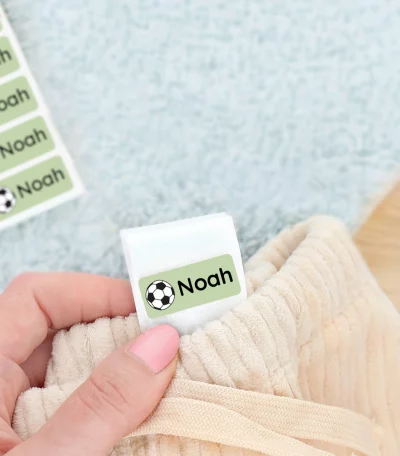 Navnelap med navnet Noah synlig i en hånd, der holder et stykke tøj med en grøn sportstema fotball. Ved siden af ligger et par grønne børnesko og et blødt tæppe. Perfekt til at personliggøre børns ejendele.