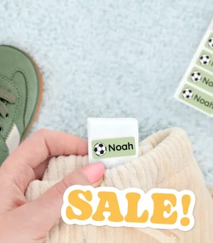 Billedet viser en hånd, der holder en blød tøjlabel med navnet Noah og et fodboldmotiv. I baggrunden ses en grøn børnesko og en stak navnelapper. Designet fremhæver muligheden for personalisering af navnelapper, ideelt til brug på tøj og sko til børn.