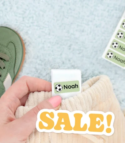 Billedet viser en hånd, der holder en blød tøjlabel med navnet Noah og et fodboldmotiv. I baggrunden ses en grøn børnesko og en stak navnelapper. Designet fremhæver muligheden for personalisering af navnelapper, ideelt til brug på tøj og sko til børn.