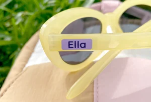 Billede viser et par gule solbriller med navnelappen Ella tydeligt synlig på siden. Baggrunden er grøn med græs, hvilket giver et friskt og sommerligt udtryk. Dette produkt er ideelt til børn, der ønsker at tilføje et personligt præg til deres accessories.