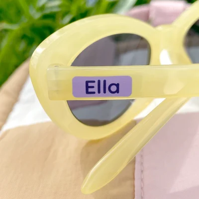 Billede viser et par gule solbriller med navnelappen Ella tydeligt synlig på siden. Baggrunden er grøn med græs, hvilket giver et friskt og sommerligt udtryk. Dette produkt er ideelt til børn, der ønsker at tilføje et personligt præg til deres accessories.