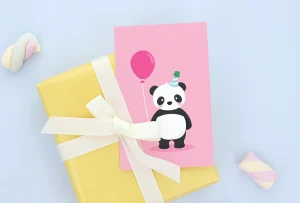 Bursdagskort med søt panda i festlig hatt, som holder en rosa ballong, plassert på en gult innpakket gave med hvit sløyfe og små pasteller i bakgrunnen. Perfekt til barnebursdager.