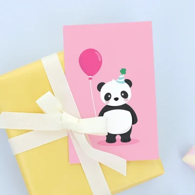Bursdagskort med søt panda i festlig hatt, som holder en rosa ballong, plassert på en gult innpakket gave med hvit sløyfe og små pasteller i bakgrunnen. Perfekt til barnebursdager.