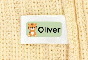 Navnelap med sød tigerillustration og navnet Oliver, placeret på et strikket, lyst baggrundsmateriale. Ideel til at personliggøre tøj og ejendele til børn.