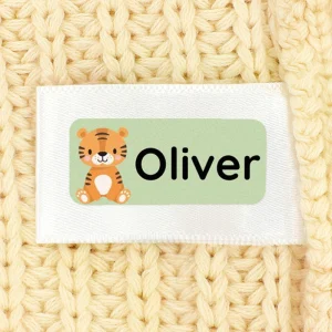 Navnelap med sød tigerillustration og navnet Oliver, placeret på et strikket, lyst baggrundsmateriale. Ideel til at personliggøre tøj og ejendele til børn.
