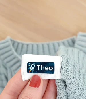 En hånd, der holder en navnelap med teksten Theo, ligger ved siden af en lyseblå striktrøje og efterårsblade på et træbord. Navnelappen er ideel til mærkning af børns tøj og ejendele.